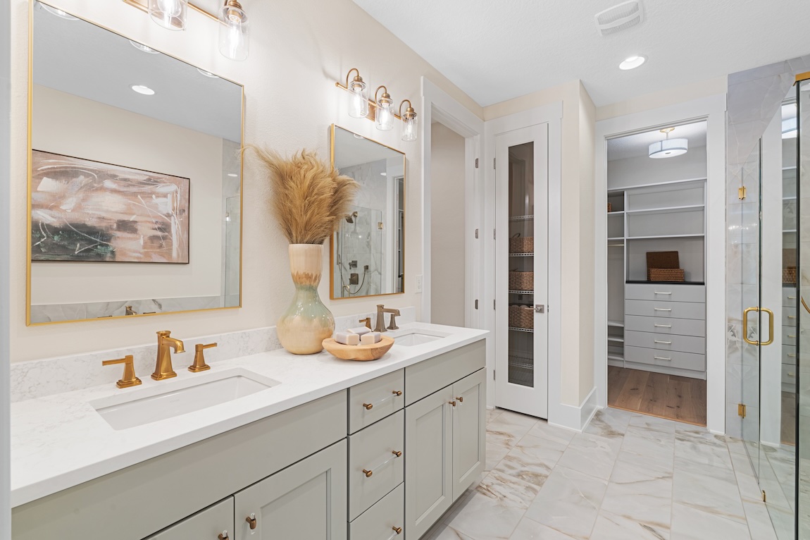 Light-filled modern bathroom with a double vanity, marble countertops, gray cabinets, and gold fixtures beside a glass shower door.