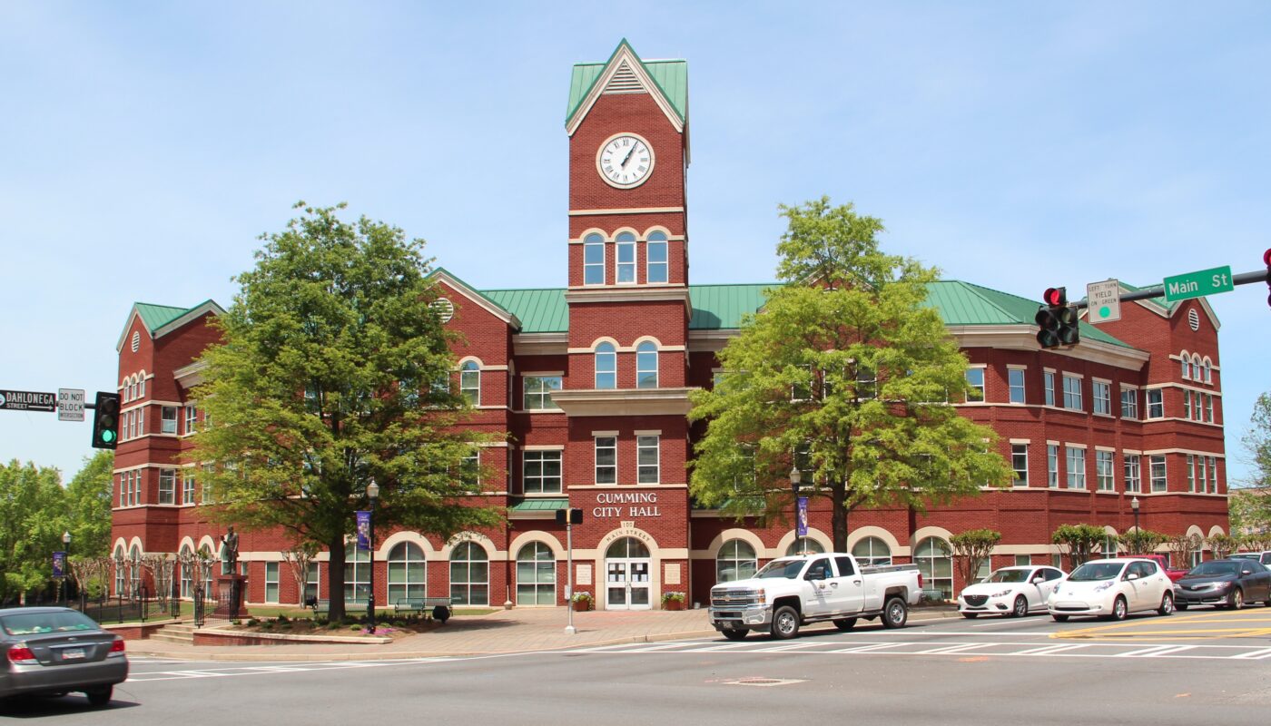 Cumming City Hall: red brick building with a green roof and clock tower at a Main Street intersection, with cars parked nearby