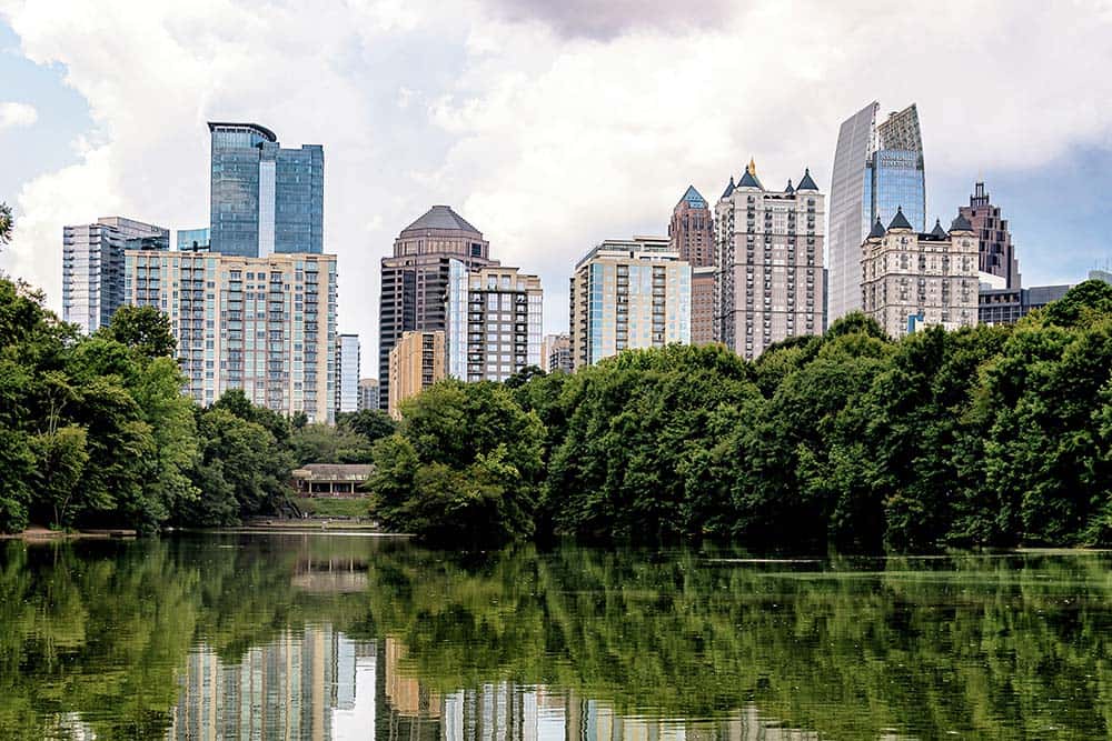 City skyline behind a dense row of trees with a calm lake in the foreground and a clear reflection of the buildings in the water.