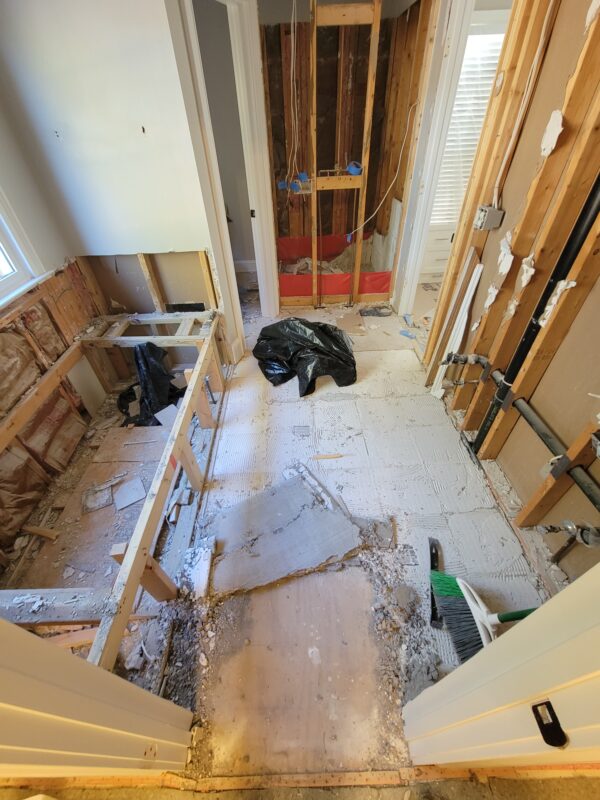 Partially demolished bathroom with exposed studs, plumbing, and debris; a black trash bag sits on the floor amid ripped drywall and construction materials. A window is visible on the left.