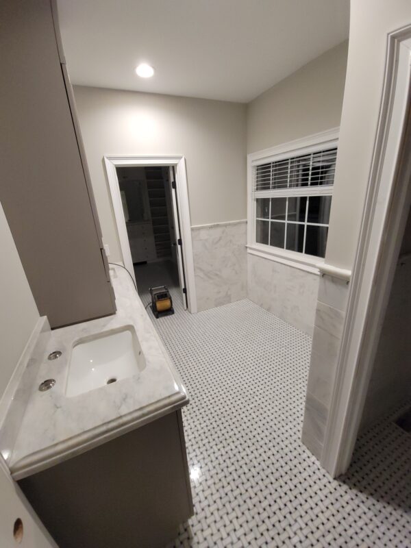 Narrow utility bathroom with marble countertop sink, gray cabinetry, and a checkered black-and-white tile floor under soft overhead lighting.