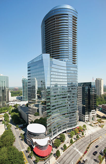Tall glass skyscraper with a curved cylindrical top in a modern office complex against a clear blue sky, seen from above a city street.