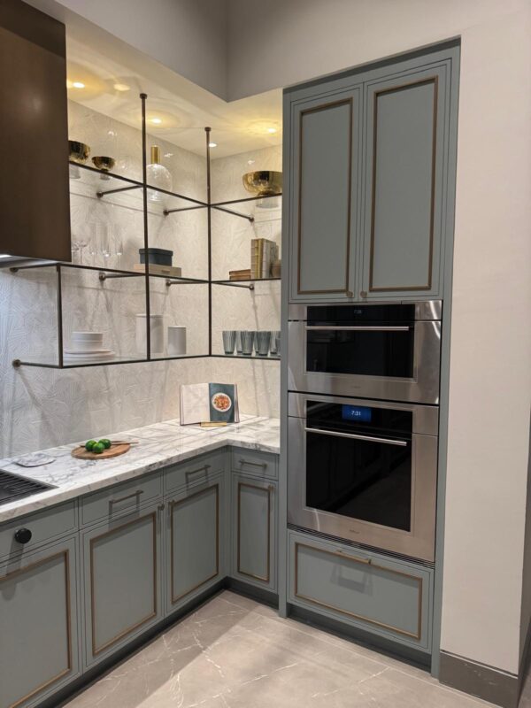 Modern grey kitchen with marble countertop, open metal shelving, and a stainless steel double oven built into a tall cabinet.