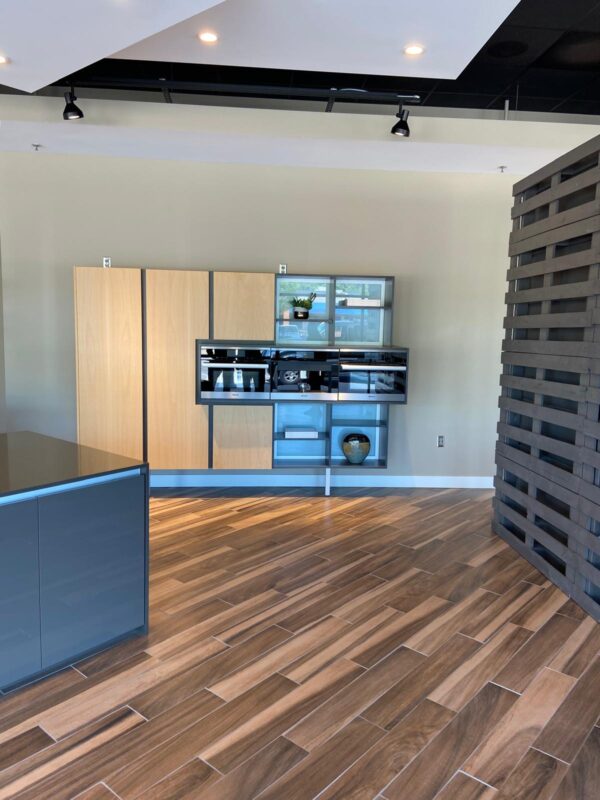 Modern kitchen display with tall wooden cabinets and built-in stainless steel ovens against a beige wall in a showroom setting.