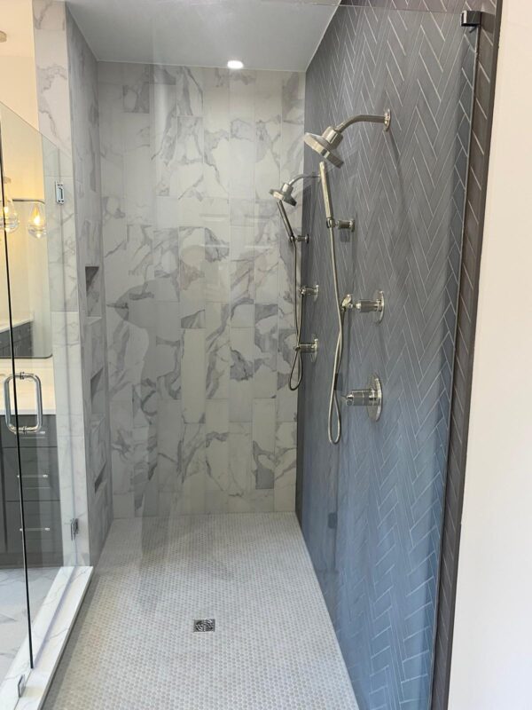 Walk-in shower with blue herringbone wall, marble tile shower area, and dual rainfall/shower heads on the right wall; glass door on the left.