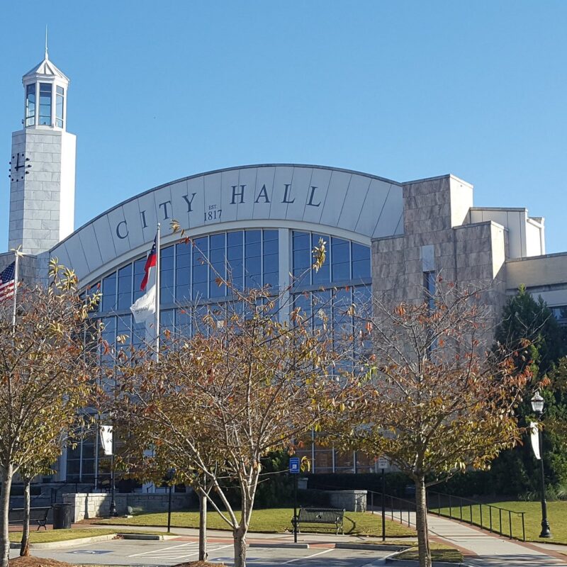 City Hall with an arched front, flags waving, and trees in the foreground on a sunny day