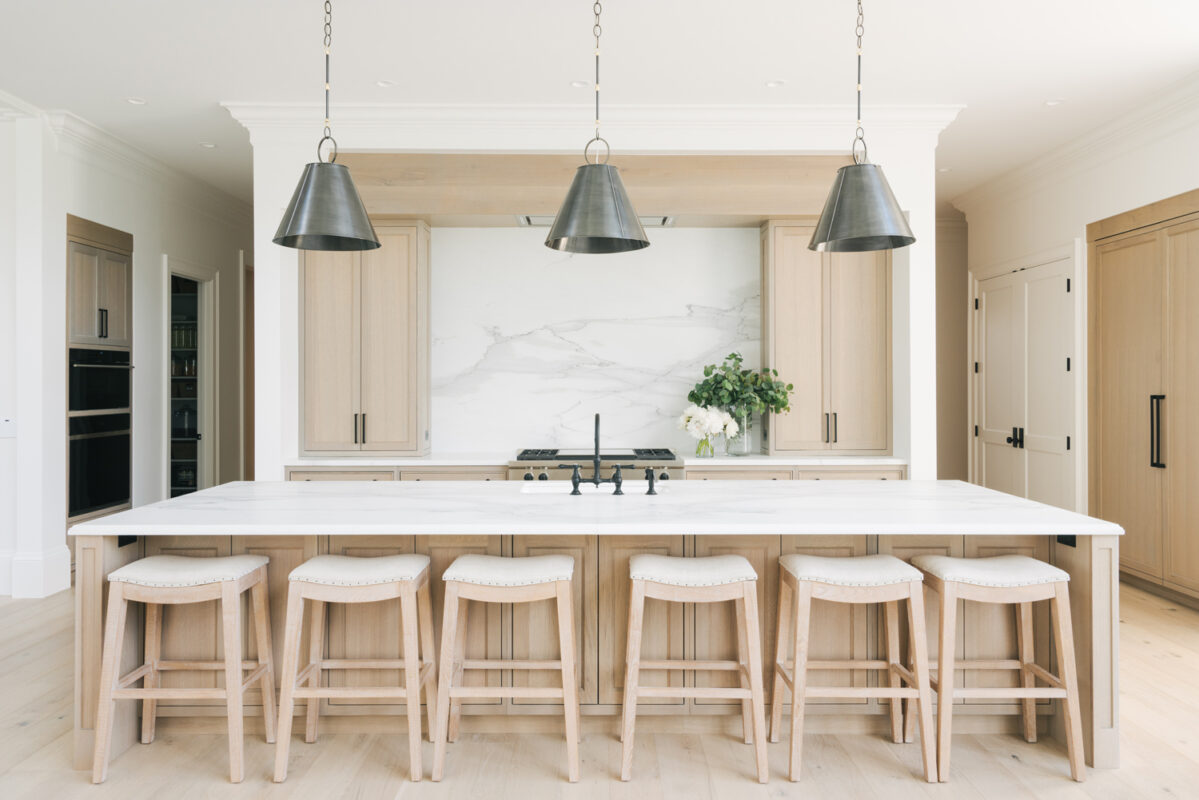 Bright modern kitchen with a large white marble island, light wood cabinets, and three metal pendant lights casting soft illumination over the counter.