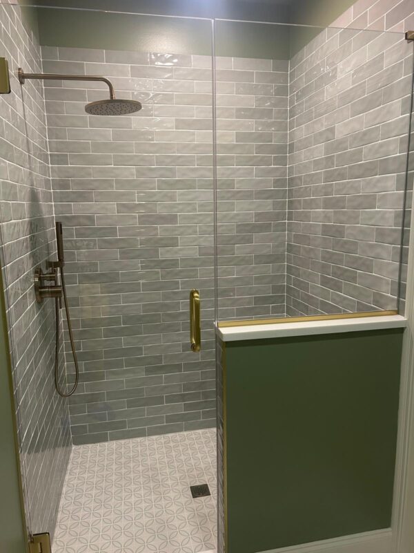 Glass-walled shower enclosure with gray subway tiles, a round rainfall shower head, and a handheld shower on the left; brass door handle and green panel divider wall beneath on the right.