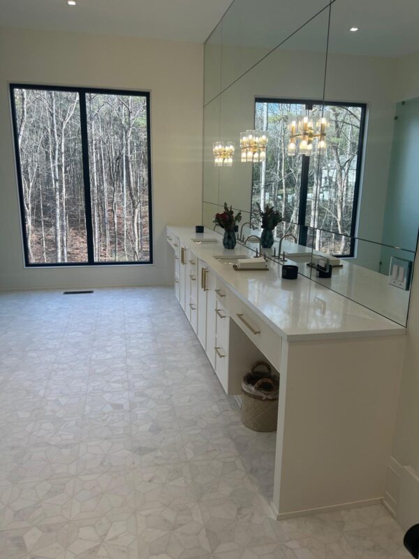Bright, modern bathroom with a long white double vanity, gold hardware, and a large mirrored wall reflecting a chandelier and forest view through two tall windows.