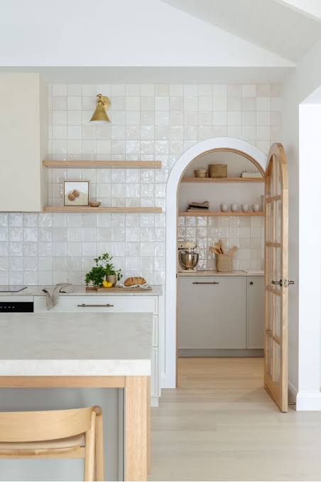 Bright kitchen with white tiled walls, open wooden shelves, and an arched doorway leading to a pantry counter with a mixer and jars.