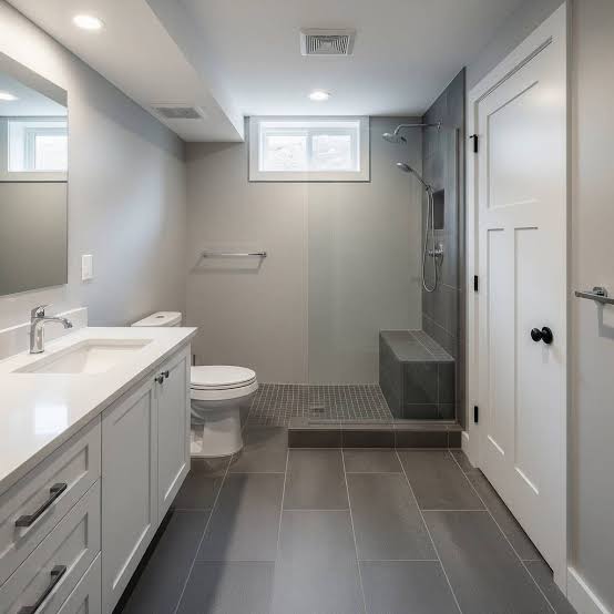 Modern bathroom with a double-sink vanity, toilet, and walk-in shower with a built-in bench; gray tile floors and walls, small high window for light.