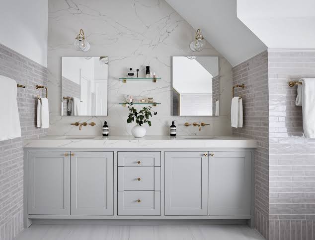 Double-sink bathroom vanity in light gray with marble countertop, brass fixtures, and two mirrors with wall-mounted towels on each side.