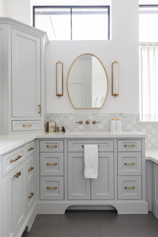Gray bathroom vanity with gold hardware, oval mirror, and two vertical gold sconces on a white wall.